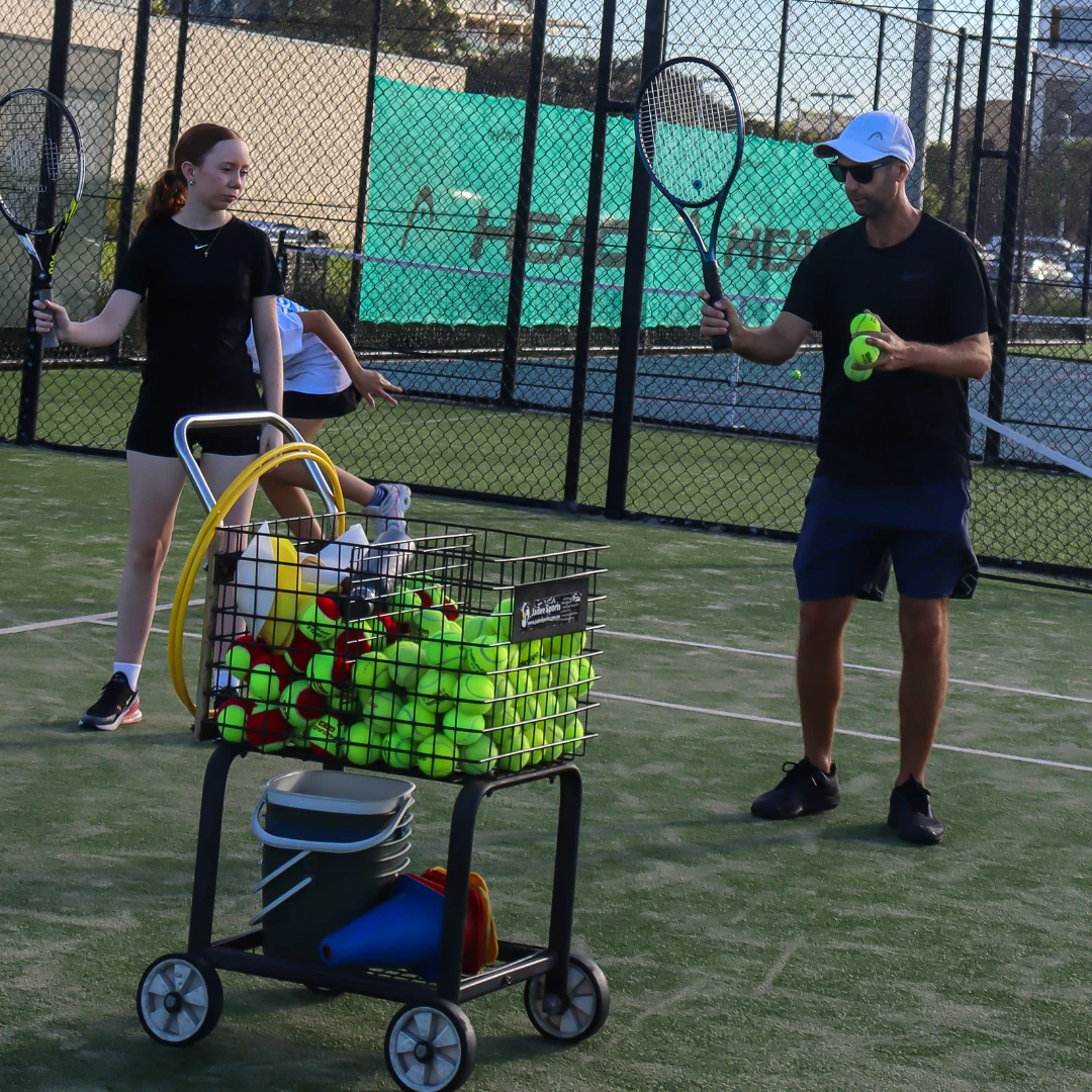 Tennis coach instructing a young player on an outdoor court, with a ball cart in the foreground.