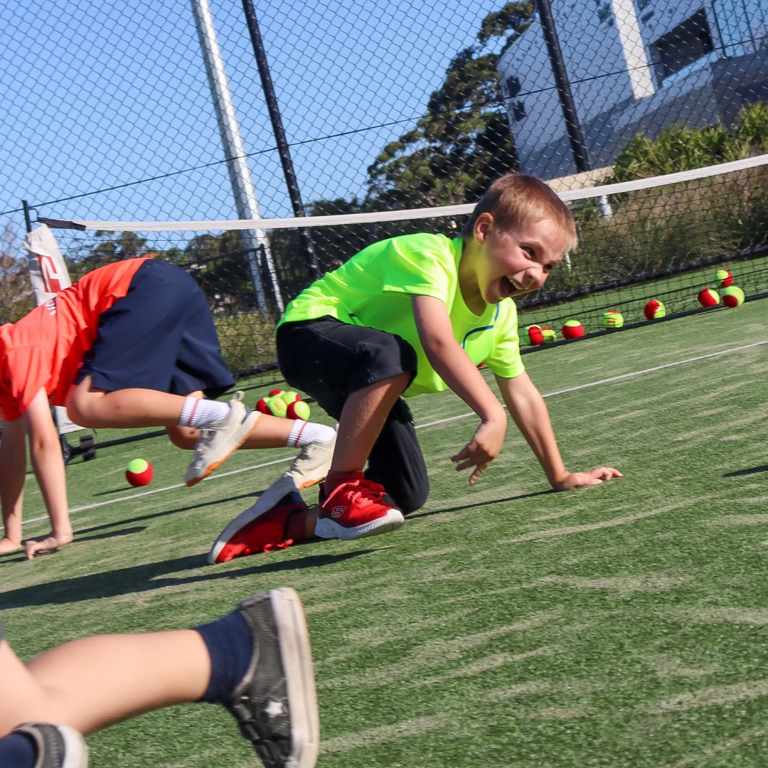 Kids laughing during a crouching ball-collecting game on a tennis court.